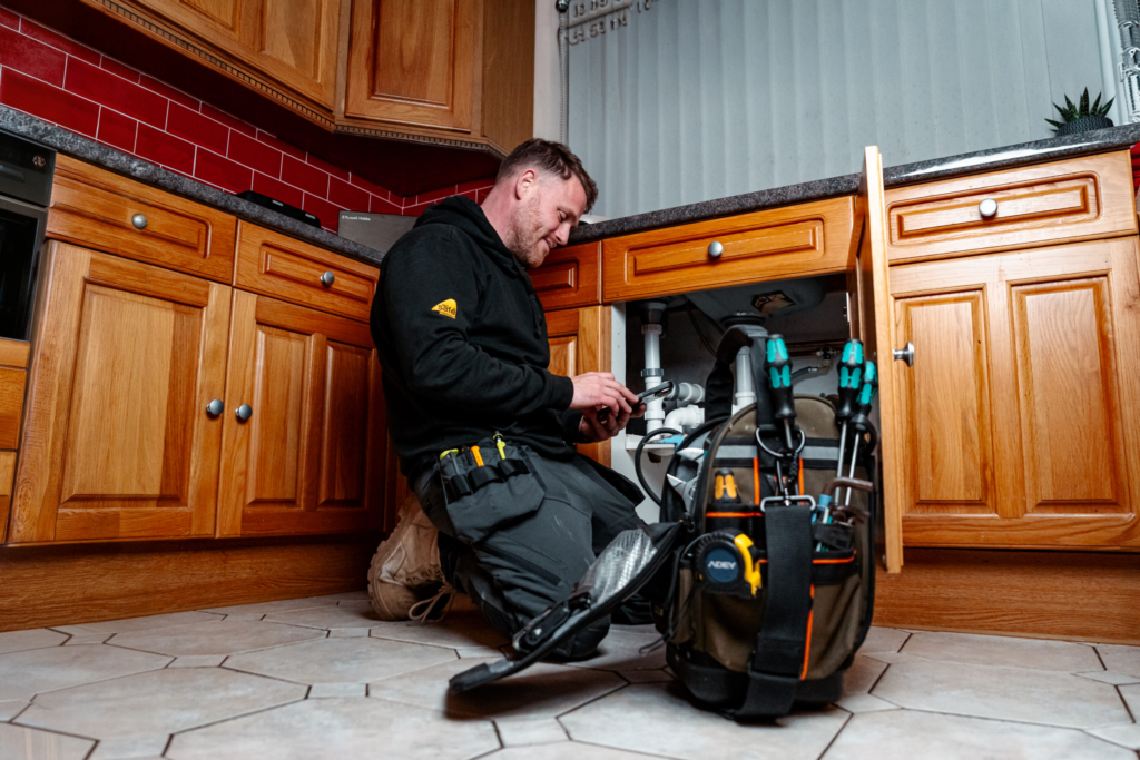 A tradesperson kneeling at a kitchen sink with his tool bag, mid-job